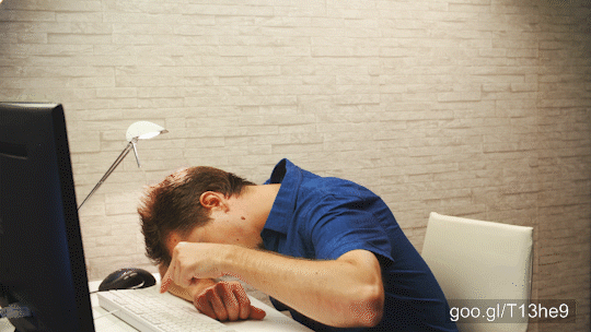 Static shot of young man in focus sleeping on the table over the arm with a screen in left side. Background office wall.