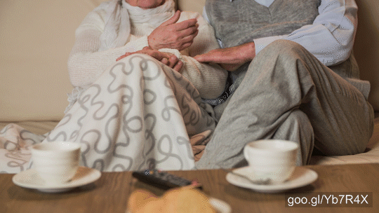 Married couple sit on sofa with tea cups on front on wooden table. Both with crossed knees and caressing male hand.