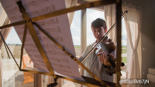 Slide shot from behind the stand with music notes to boy playing on violin at home with big bright windows in background.